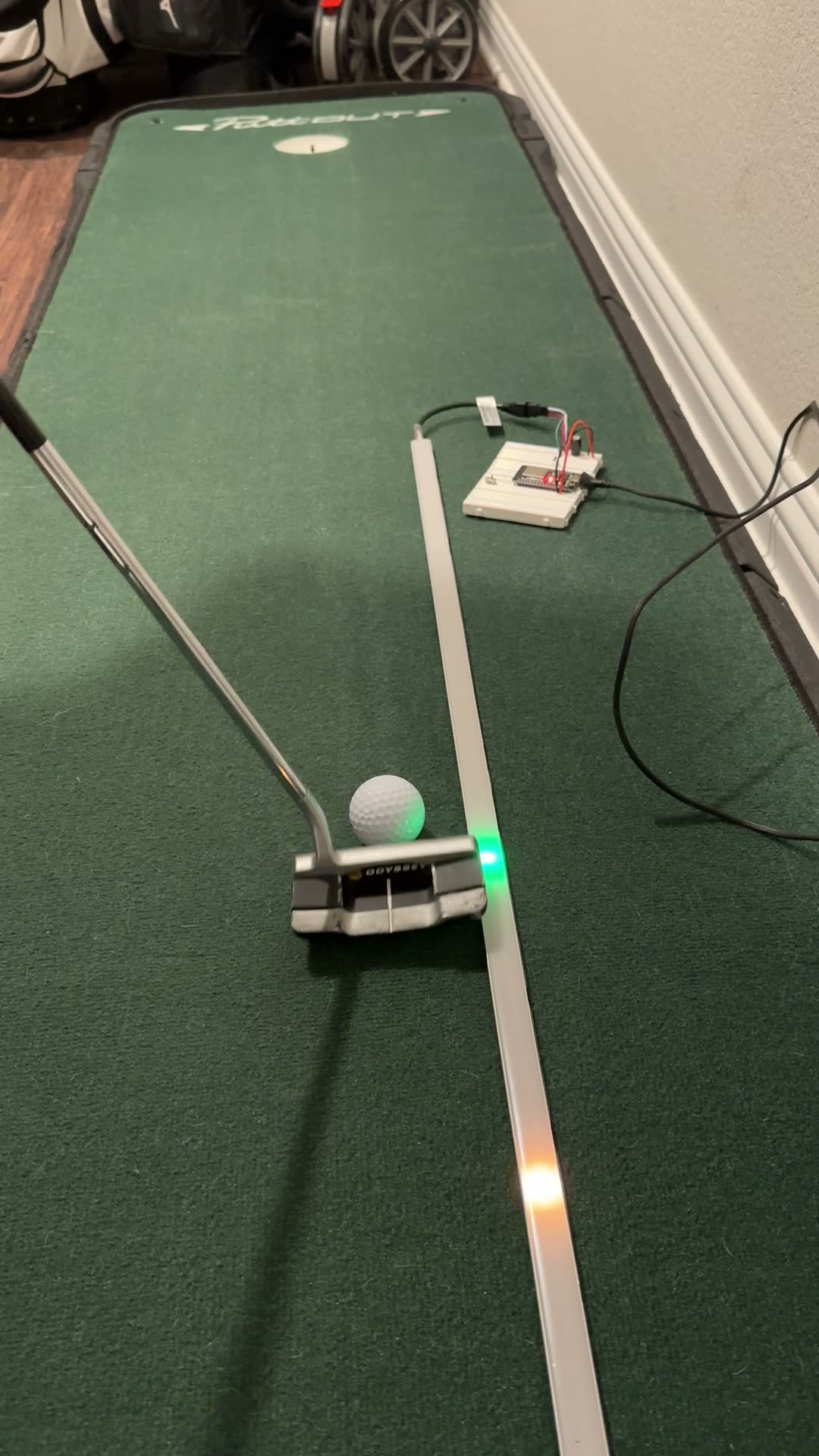 OSSTempo on a putting mat: a putter and ball beside a WS2812B LED strip showing a green impact dot and a white comet, driven by an ESP32 on a breadboard